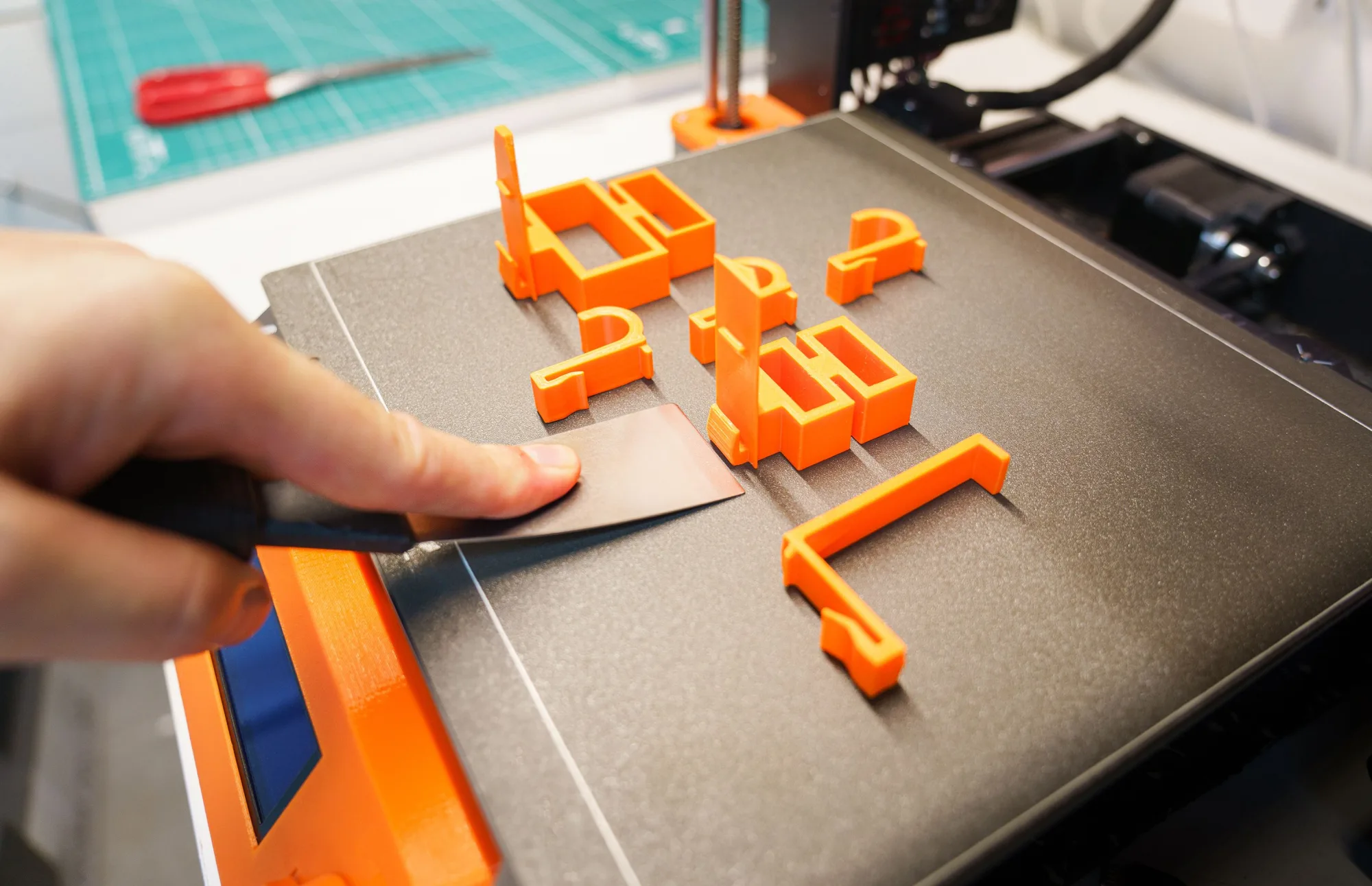 Eine Hand löst orangefarbene Bauteile vom Druckbett eines 3D-Druckers.