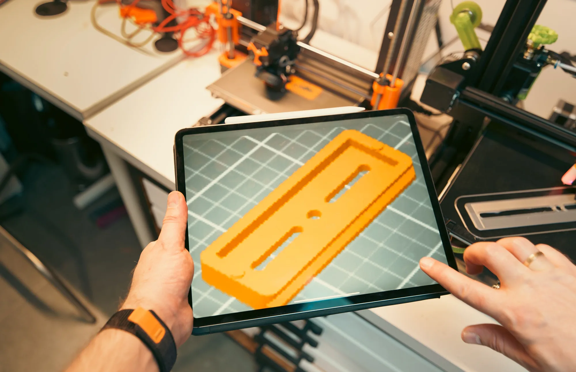 Eine Person betrachtet ein digitales 3D-Modell auf einem Tablet neben mehreren 3D-Druckern.