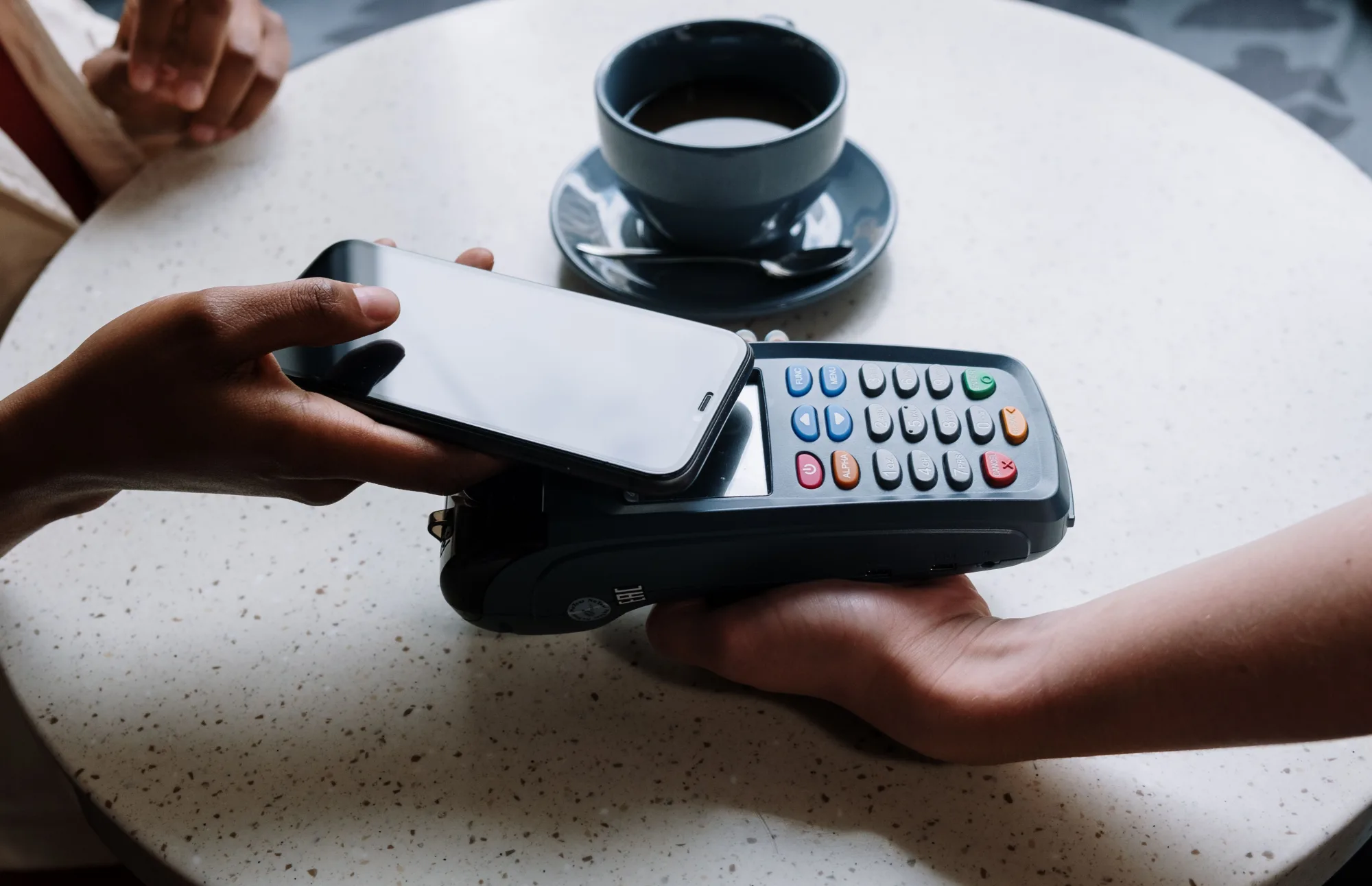 Hände halten ein Smartphone über ein Kartenterminal auf einem Cafétisch