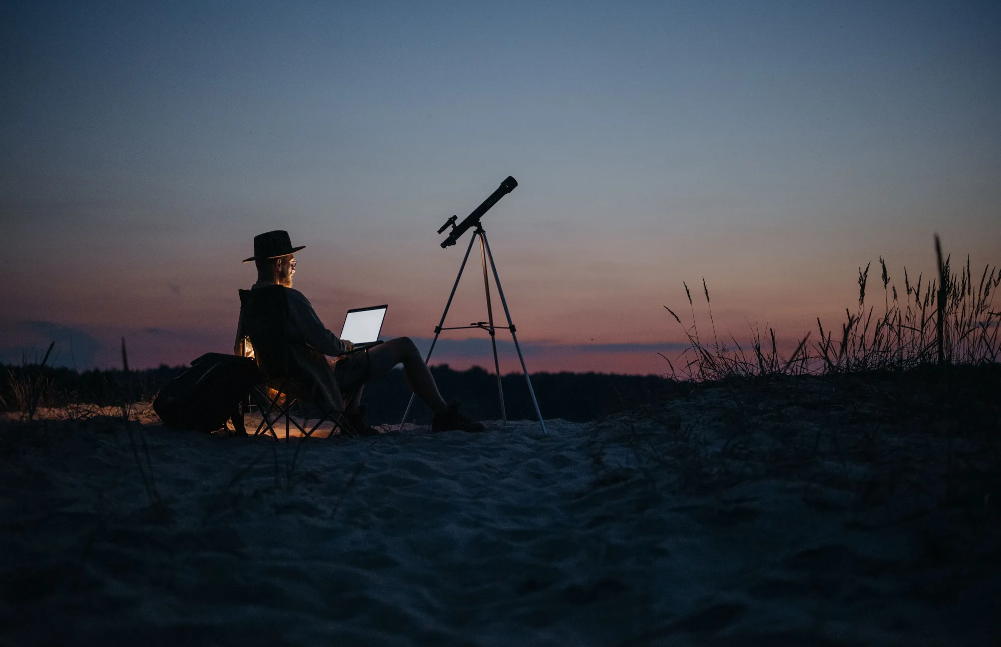 Eine Person arbeitet bei Dämmerung neben einem Teleskop am Laptop.